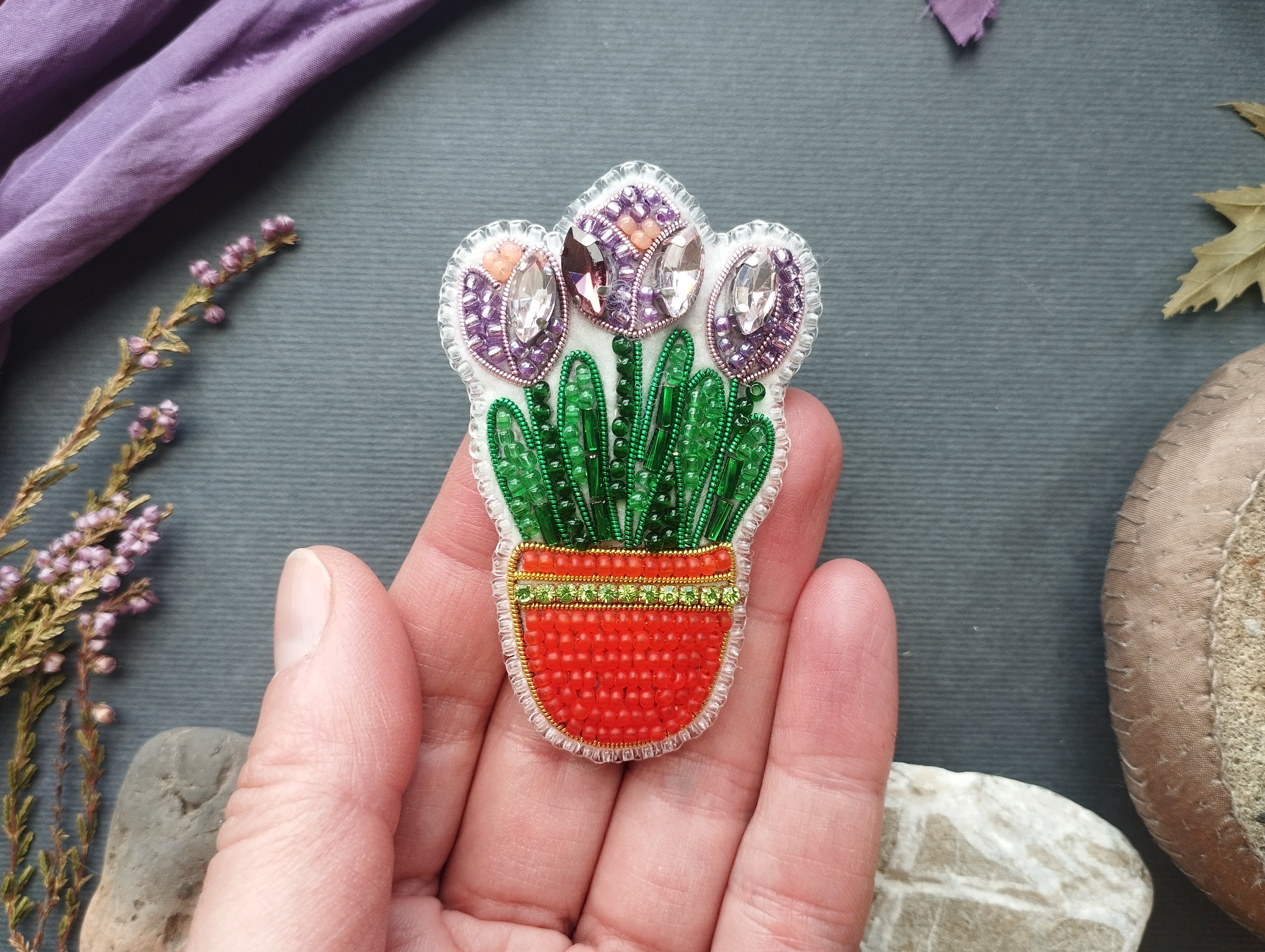 A hand holding a decorative pin featuring a plant with purple flowers and green leaves, placed on a gray surface with various natural elements such as leaves and flowers.